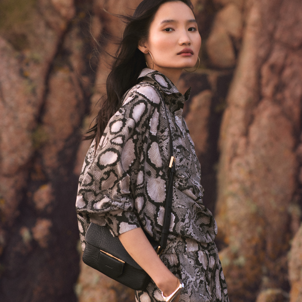 Model wearing a snake skin shirt and trouser set with a black handbag sold at AllSaints