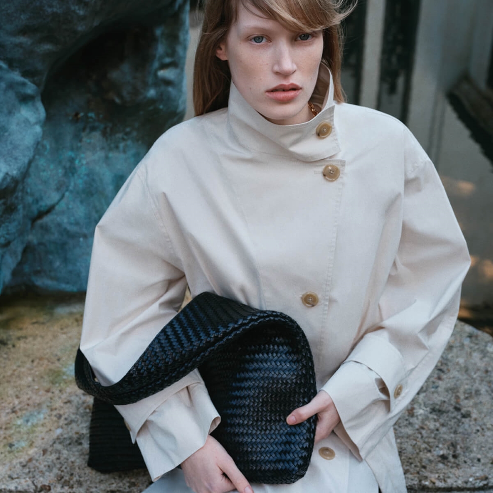 Model wearing a high neck off white trench coat holding a black hobo bag sold at Mango